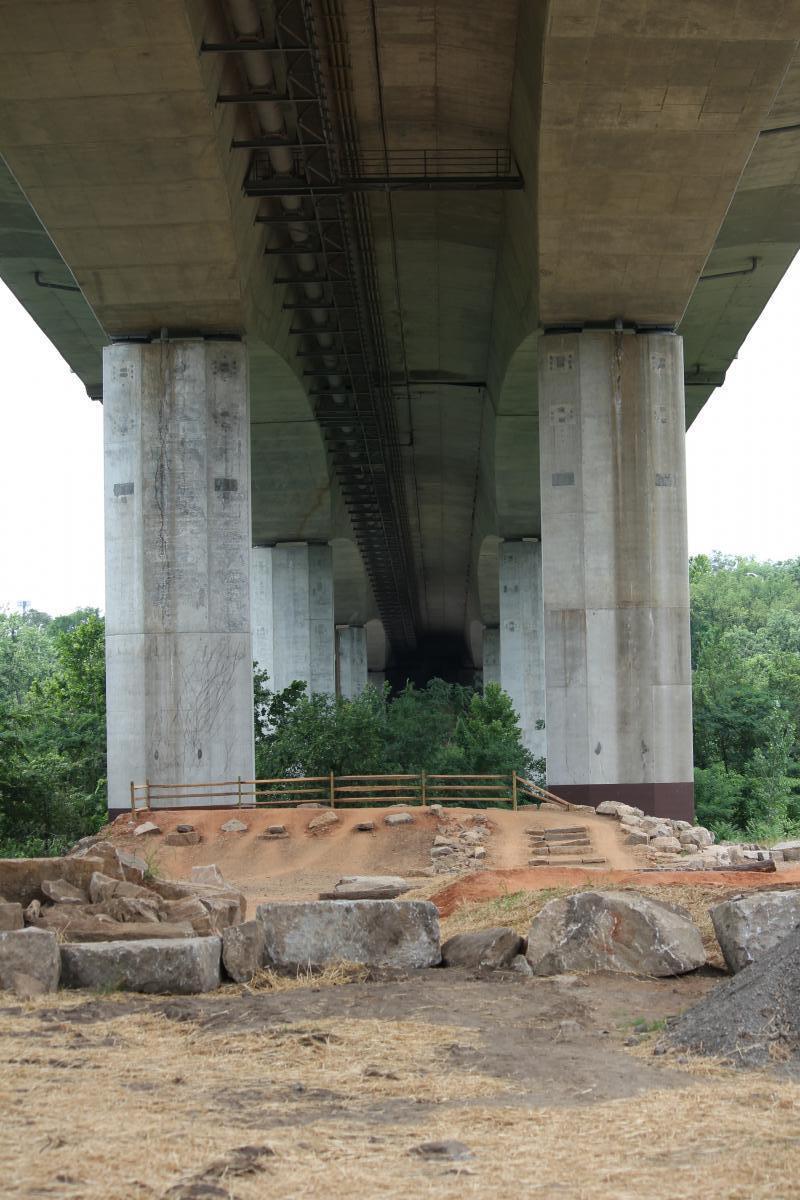 Alt text: "View from underneath a large concrete bridge, showing thick supporting pillars and a dirt area with rocks and a wooden fence in the foreground, surrounded by green trees." Belle Isle mountain bike trail.
