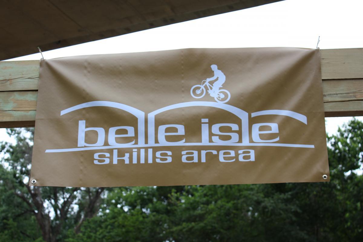 A banner for "Belle Isle Skills Area" featuring white text on a brown background, with a silhouette of a cyclist performing a trick. It is displayed on a wooden structure, surrounded by greenery. Belle Isle mountain bike trail.