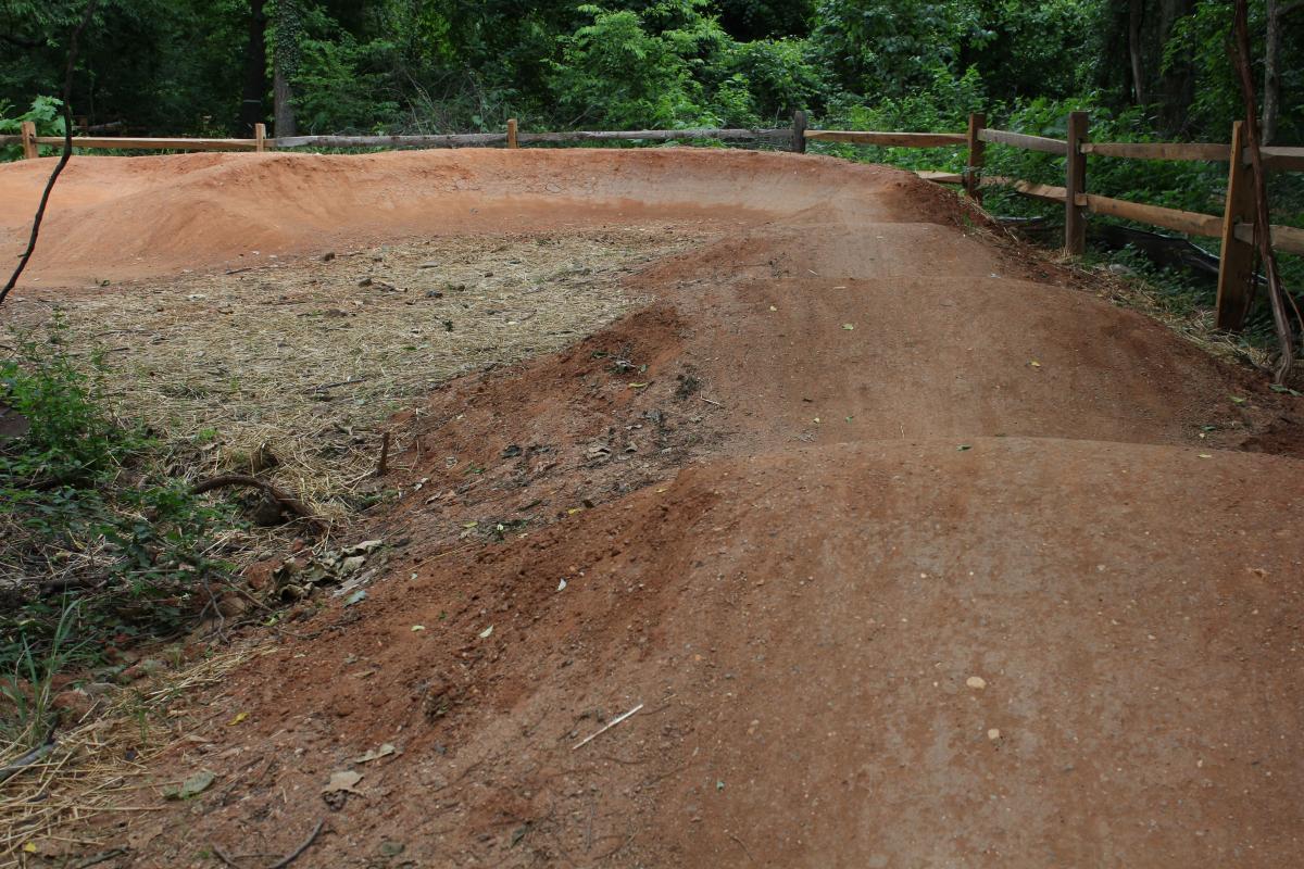 A dirt bike track featuring a winding path with smooth curves, surrounded by greenery and a wooden fence. The track is composed of reddish-brown dirt, with some areas showing straw and small vegetation nearby. Belle Isle mountain bike trail.