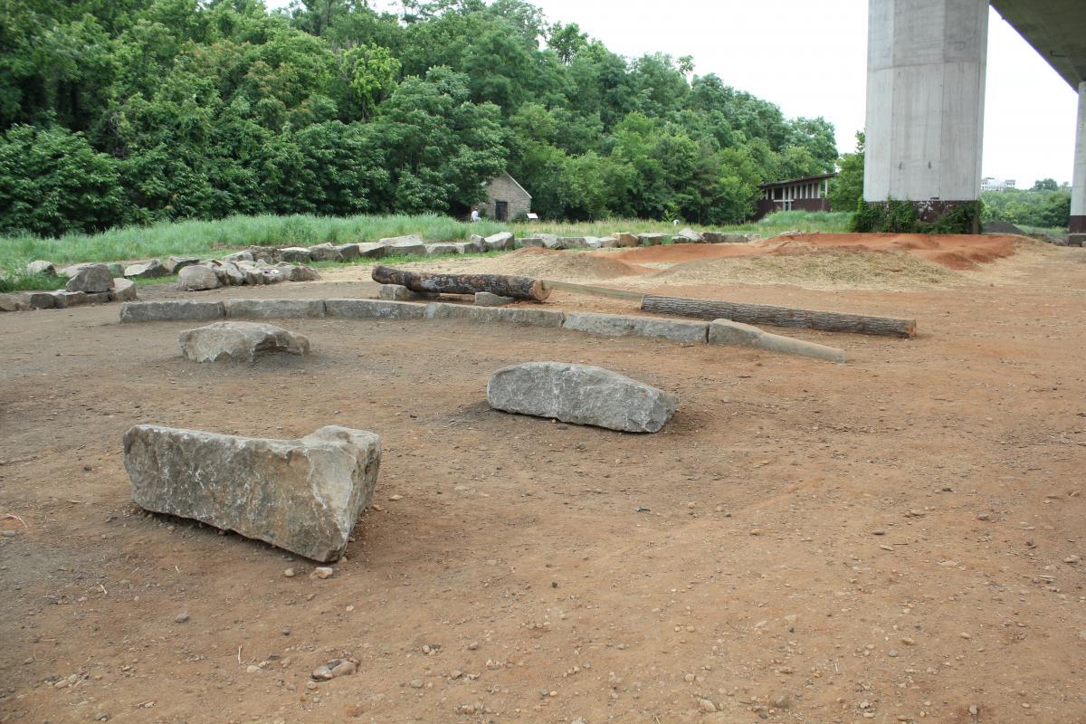 Excavation site featuring scattered stones and logs under a concrete structure, with a backdrop of green trees and grass. Belle Isle mountain bike trail.