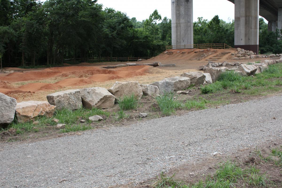 A dirt bike track under a bridge, featuring gravel pathways, earthen ramps, and large rocks lining the edge. The area is surrounded by trees and greenery, with a wood fence in the background. Belle Isle mountain bike trail.
