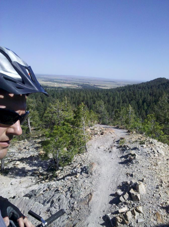 Mountain biker standing on a rocky trail with a scenic view of green forests and distant plains under a clear blue sky. Cheyenne Mountain State Park mountain bike trail.