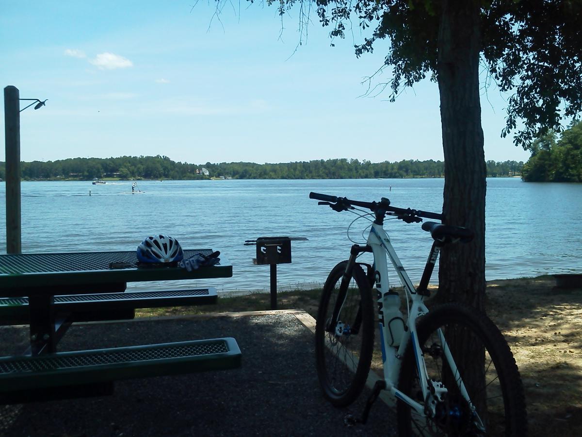 A serene lakeside scene featuring a white mountain bike resting against a picnic table under a tree. Nearby, a bicycle helmet and gloves are placed on the table. The calm lake is visible in the background, with a paddleboarder and a boat in the distance, framed by lush greenery and a clear blue sky. Arrowhead Park mountain bike trail.