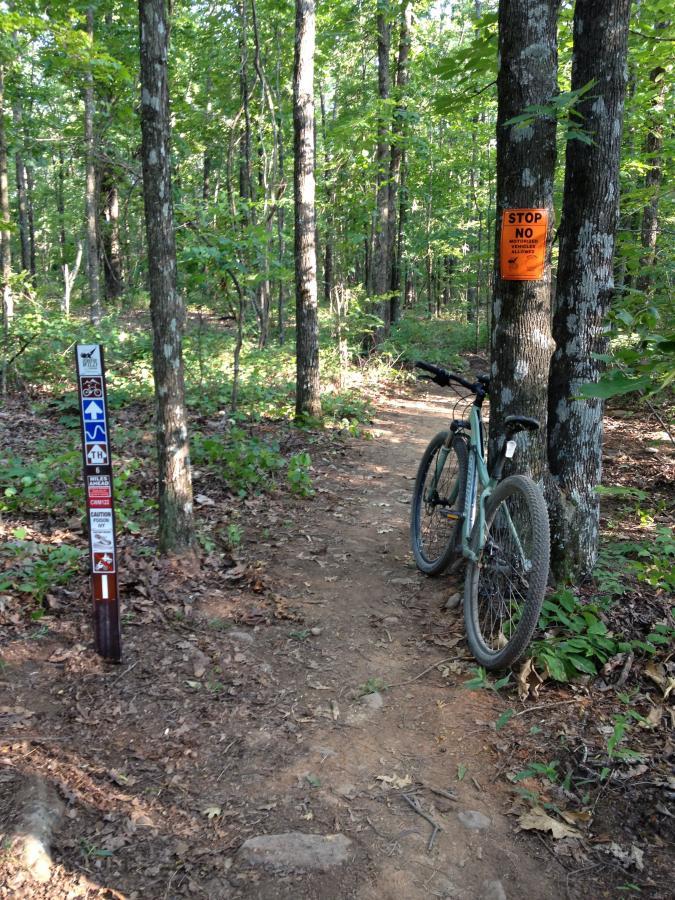 A mountain bike parked beside a dirt trail in a wooded area, with a trail signpost displaying various biking symbols and a caution sign reading "STOP NO." Tall trees and green foliage surround the path, indicating a natural outdoor setting. Coldwater Mountain mountain bike trail.