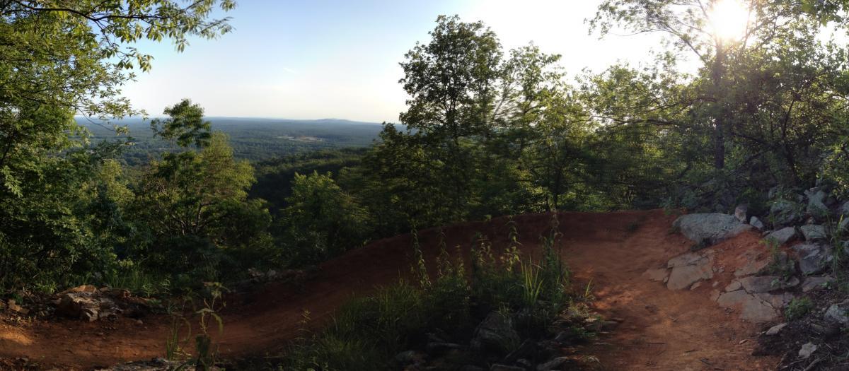 Panoramic view of a forested landscape with rolling hills in the distance, framed by lush green trees and a winding dirt path. The sun is setting, casting a warm glow on the scene. Rocks and tall grasses are visible along the path, indicating a natural hiking or biking trail. Coldwater Mountain mountain bike trail.