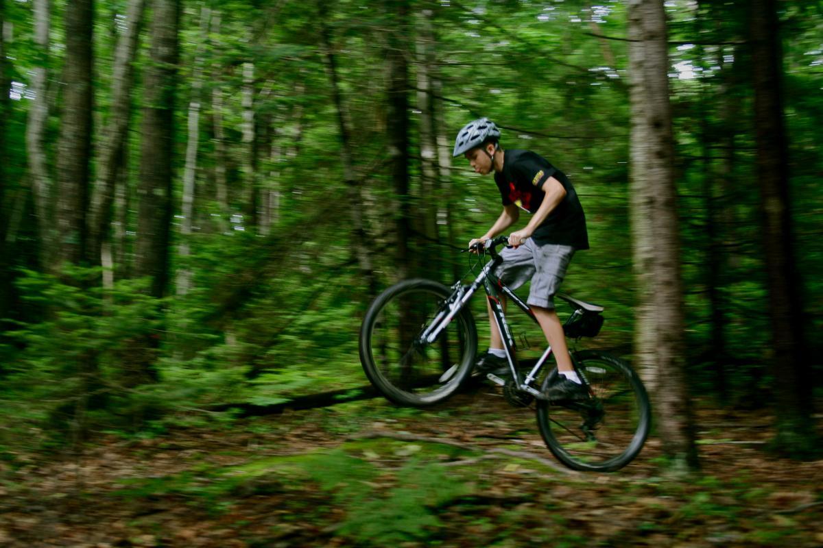 A young cyclist in a black helmet and a black T-shirt is mid-air while jumping over a log on a mountain bike in a lush forest setting. The scene captures motion, with trees and greenery blurred in the background, emphasizing the action. Glenden
