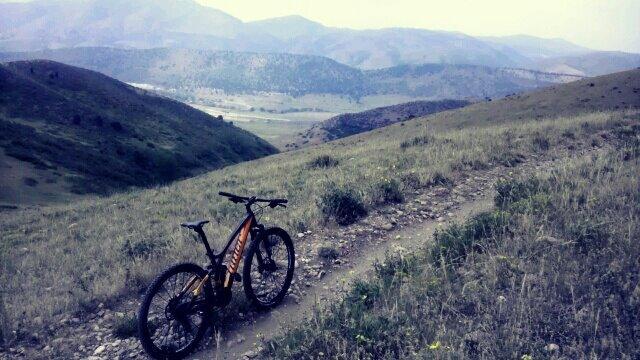 A mountain bike resting on a dirt trail, surrounded by lush green grass and rolling hills, with a backdrop of distant mountains under a cloudy sky. Green Mountain mountain bike trail.