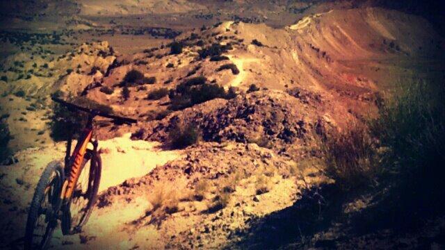 A mountain bike resting on rocky terrain, overlooking a vast, rugged landscape with hills and valleys in the background. The scene showcases a mix of earth tones and natural vegetation, suggesting an outdoor adventure environment. White Ridge Bike Trails mountain bike trail.