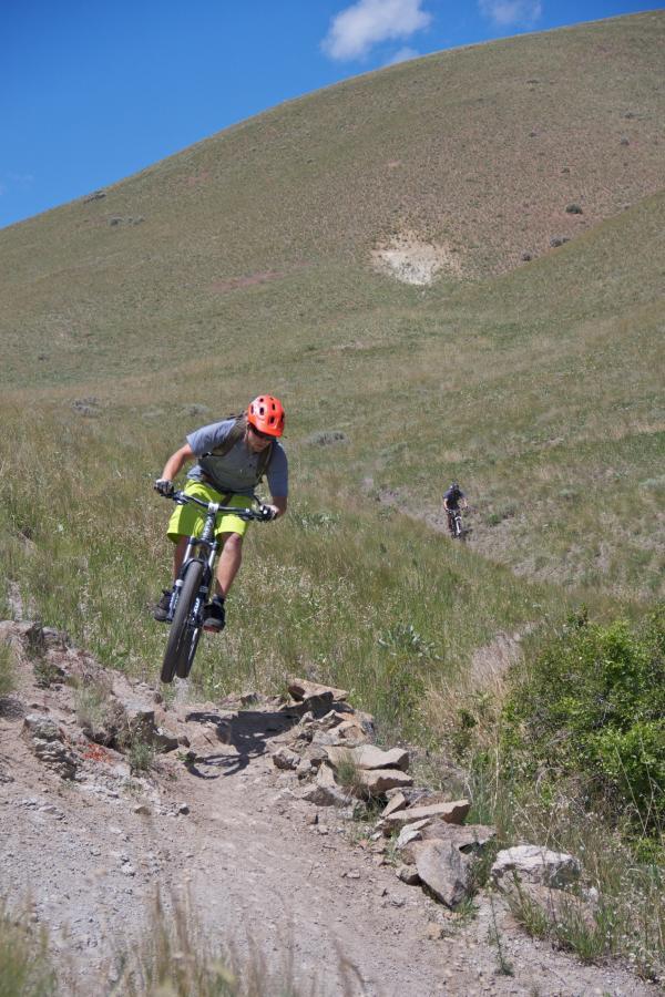 A mountain biker in a bright orange helmet and gray shirt performs a jump on a rocky trail, surrounded by grassy hills under a blue sky with a few clouds. In the background, another cyclist rides along the path. Wagonhammer Trail System mountain bike trail.