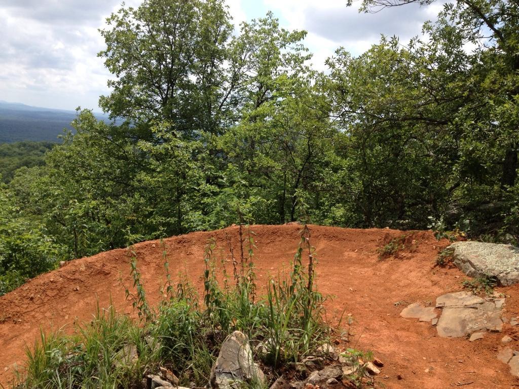 A dirt trail curving at the edge of a forested overlook, surrounded by green trees and shrubs, with a distant view of rolling hills under a cloudy sky. Coldwater Mountain mountain bike trail.