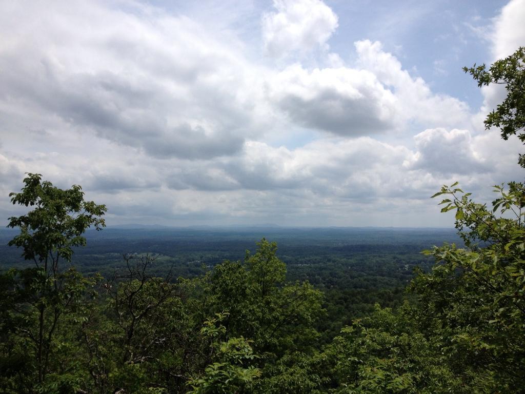 A scenic view of a lush green valley surrounded by distant mountains under a partly cloudy sky. In the foreground, trees frame the view, highlighting the natural landscape. Coldwater Mountain mountain bike trail.