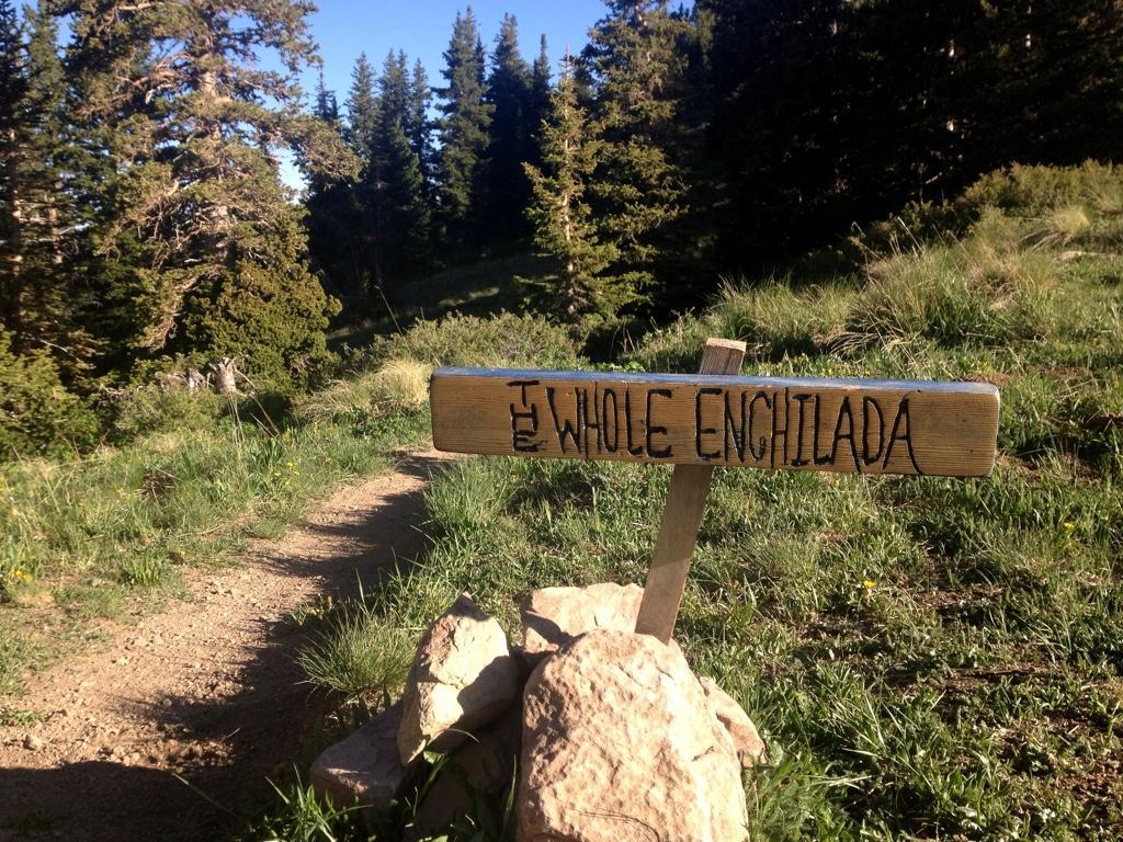 A wooden trail sign reading "The Whole Enchilada," positioned on a dirt path surrounded by grassy terrain and evergreen trees in the background. The Whole Enchilada mountain bike trail.