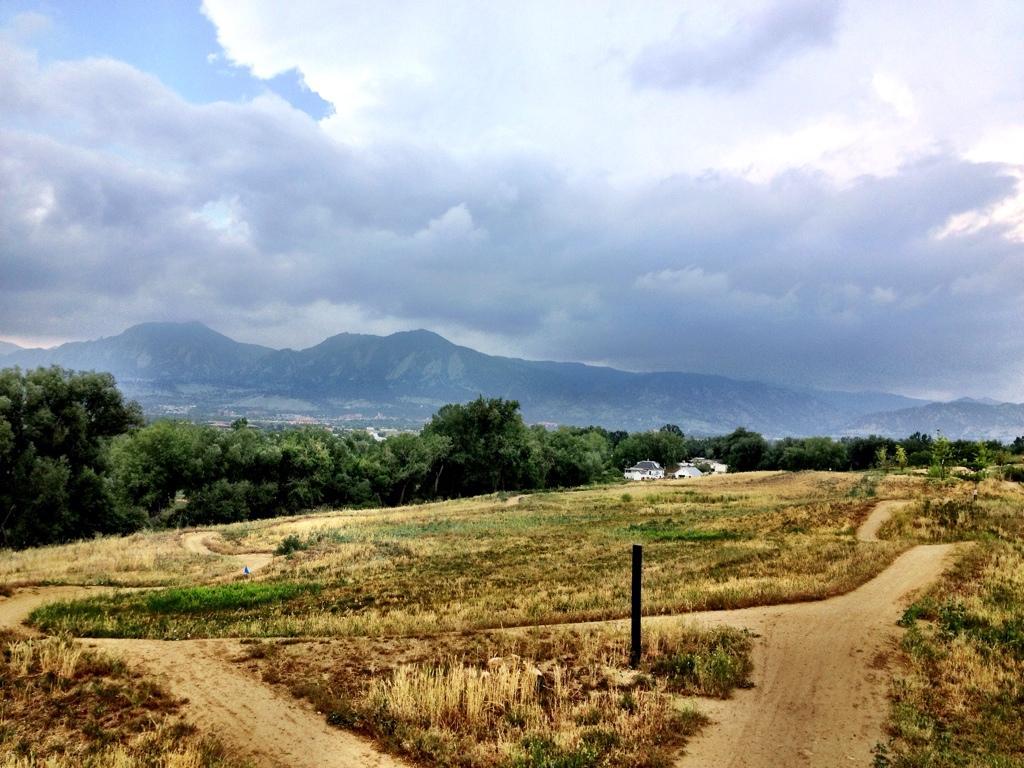 A panoramic view of a landscape featuring rolling hills and distant mountains under a cloudy sky. A winding dirt path meanders through a grassy field, with patches of green and dry grass visible. Trees line the sides, and a few houses are seen in the background, creating a serene outdoor scene. Valmont Bike Park mountain bike trail.