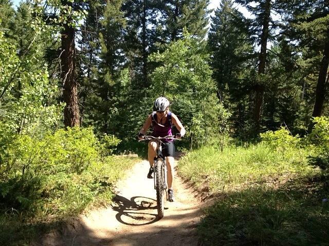 A person riding a mountain bike along a sunlit dirt trail surrounded by tall trees and greenery. The cyclist is focused and wearing a helmet, showcasing an active outdoor lifestyle. Meyer Ranch Park mountain bike trail.