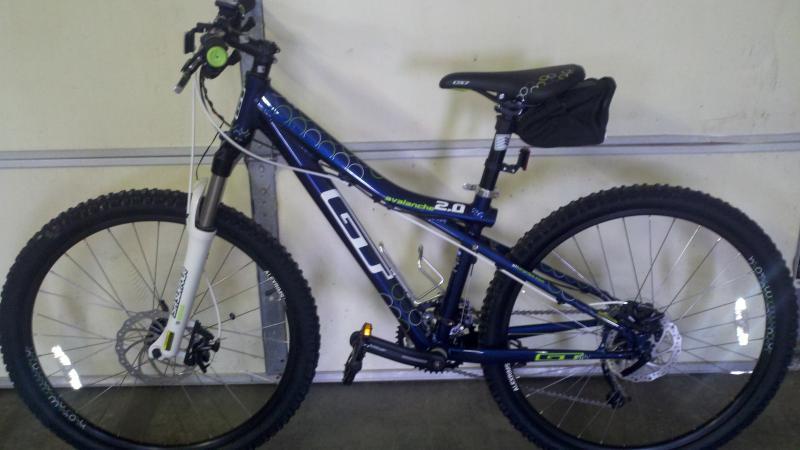 GT Avalanche 2.0: A blue and black mountain bike leaning against a wall, featuring large tires, a front suspension fork, and a small saddlebag attached to the seat. The bike's frame displays the model name "Avalanche 2.0."