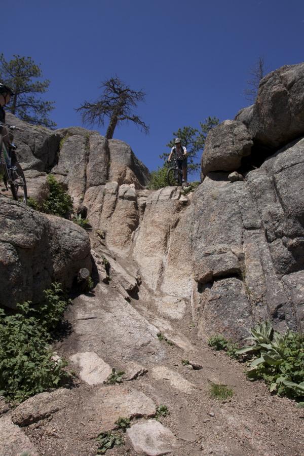 Two mountain bikers navigate a rocky terrain under a clear blue sky. The scene features steep rock formations and sparse vegetation, creating a rugged outdoor environment. One biker is positioned at the top of a steep drop, while the other is further down the slope, showcasing the challenges of the trail. Blackjack / Raspberry Ridge mountain bike trail.