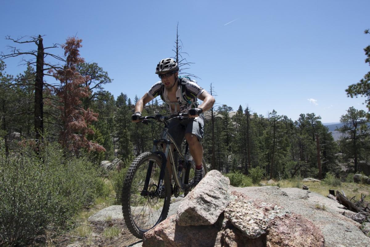 A mountain biker navigating rocky terrain in a forested area. The cyclist is wearing a helmet and a cycling jersey, and appears to be focused as they ride over a boulder surrounded by trees and a clear blue sky. Buffalo Creek mountain bike trail.