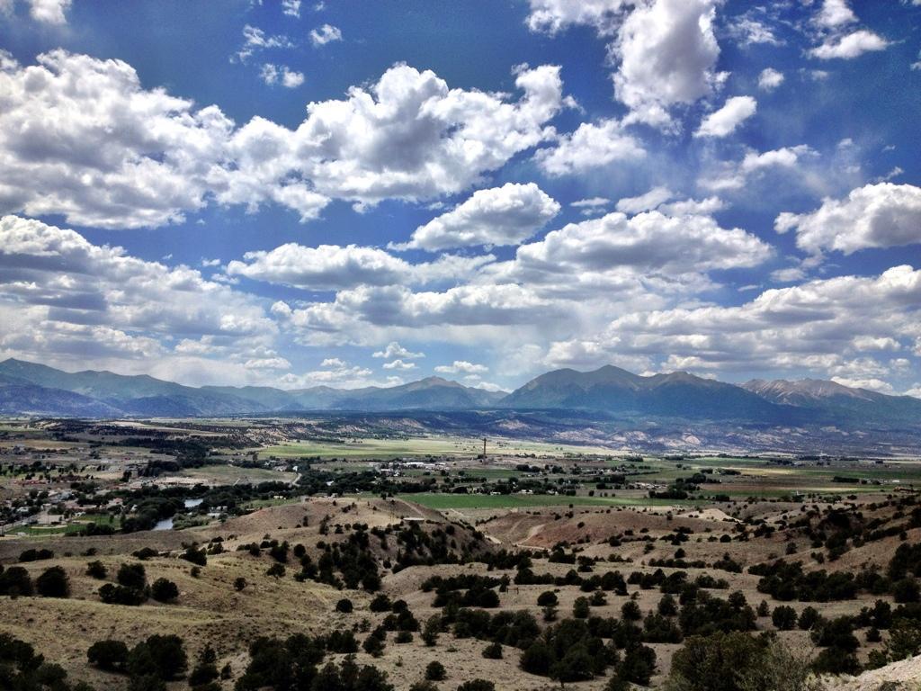 A panoramic view of a mountainous landscape under a partly cloudy sky. Rolling hills and green valleys are visible in the foreground, with a network of homes and roads scattered throughout the terrain. The distant mountains rise majestically against the backdrop of billowy clouds. North Backbone mountain bike trail.