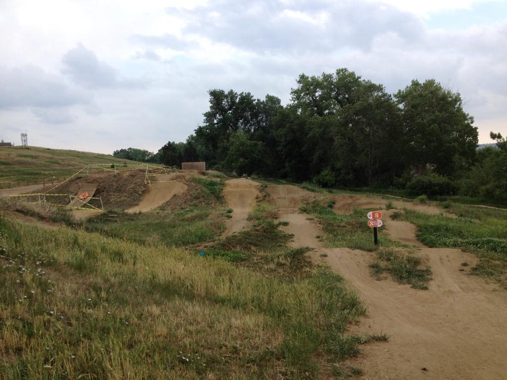 A dirt bike track with multiple jumps and berms surrounded by grassy areas, trees in the background, and cloudy skies overhead. A sign with numbers indicates the track layout. Valmont Bike Park mountain bike trail.