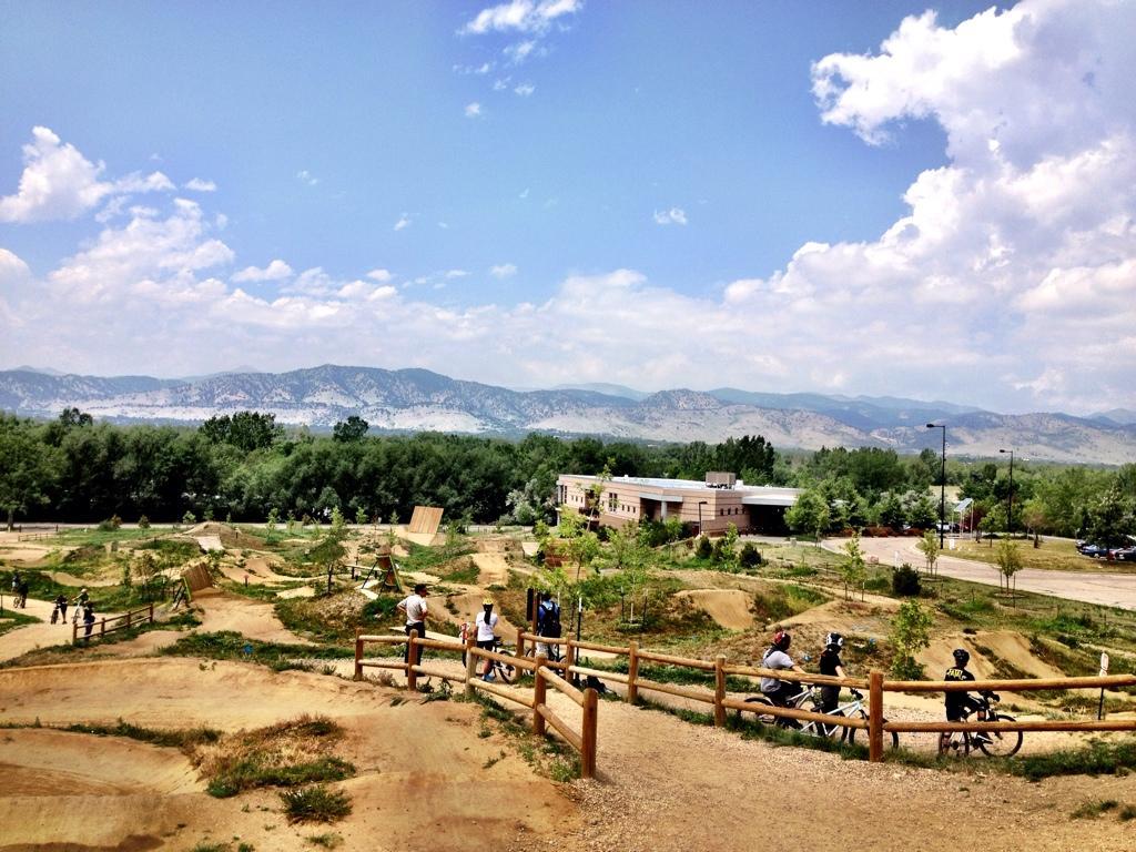 A scenic view of a bike park featuring dirt jumps and trails, surrounded by lush greenery and distant mountains under a partly cloudy sky. A few cyclists are seen navigating the trails, and a modern building is visible in the background. Valmont Bike Park mountain bike trail.