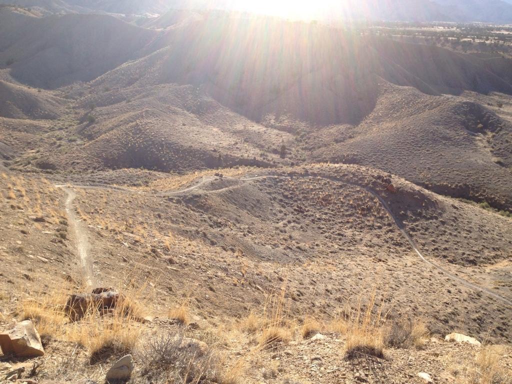A picturesque view of rolling hills and rugged terrain under bright sunlight, showcasing a winding dirt trail along the hillside. The landscape features sparse vegetation and dry grasses, typical of a mountainous region, with the sun casting a warm glow over the scene. Zippety Do Dah mountain bike trail.