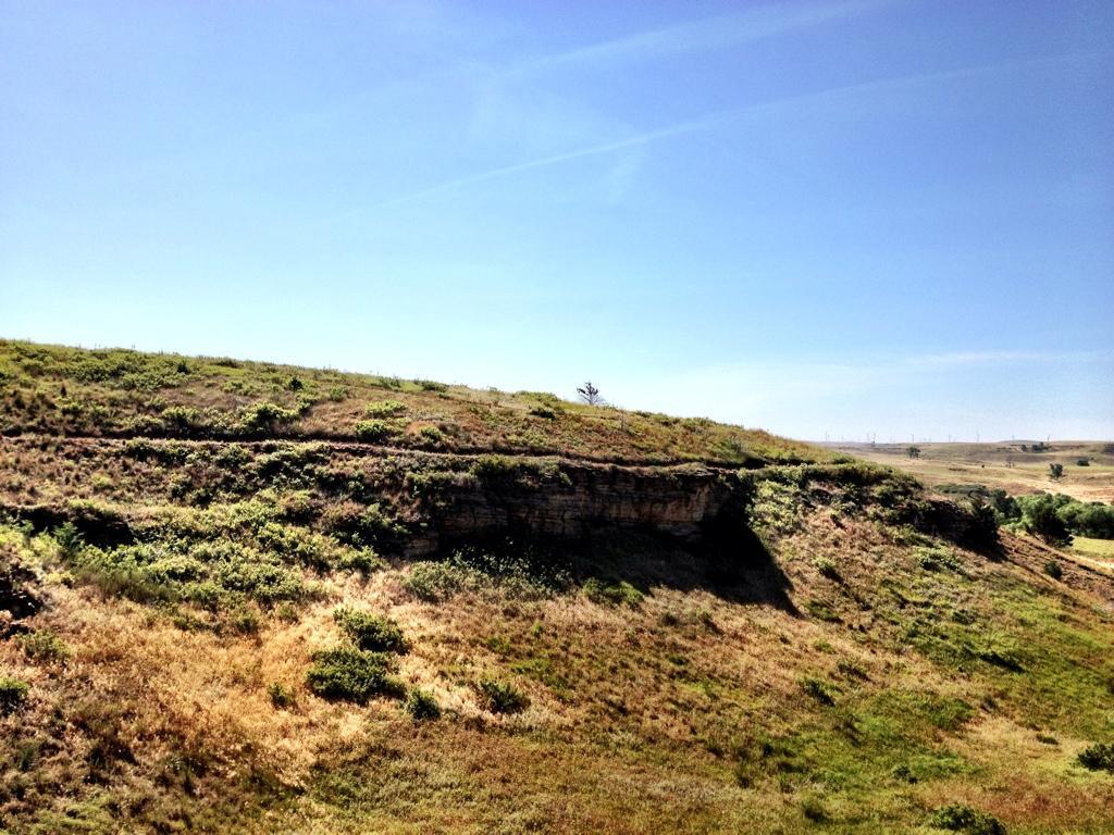 A scenic landscape featuring a gently sloping hill with green vegetation and patches of dry grass under a clear blue sky. The hill's edge reveals rocky layers, and in the distance, a flat, expansive terrain extends, hinting at distant hills and wind turbines. Switchgrass mountain bike trail.