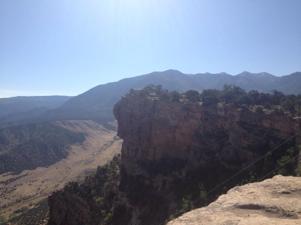 A panoramic view of a rocky cliff edge overlooking a vast mountainous landscape, with rolling hills and a blue sky. The scene captures the natural beauty of the terrain, featuring rugged rock formations and sparse vegetation in the foreground, while tall mountains rise in the background under bright sunlight. UPS And LPS mountain bike trail.