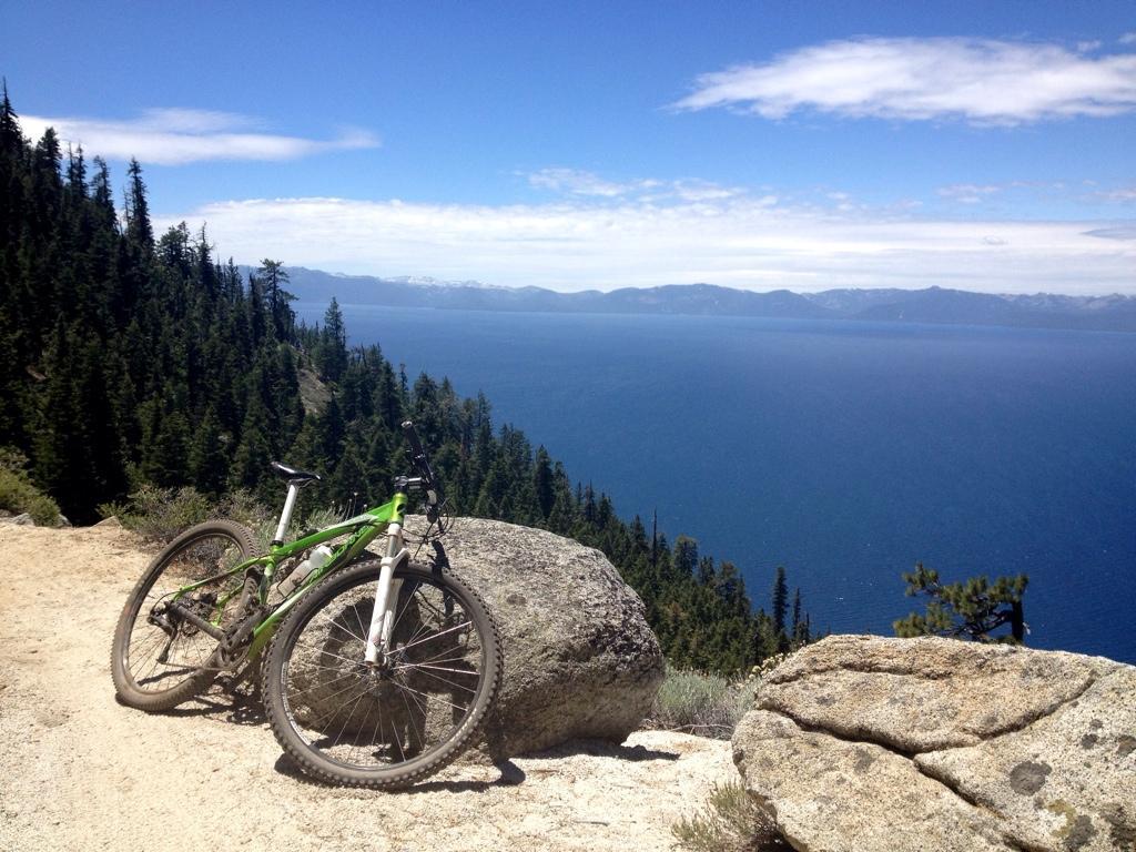 A green mountain bike resting against a large rock with a scenic view of a blue lake and distant mountains under a partly cloudy sky. Pine trees are visible in the foreground, indicating a forested area surrounding the lake. Flume Trail mountain bike trail.