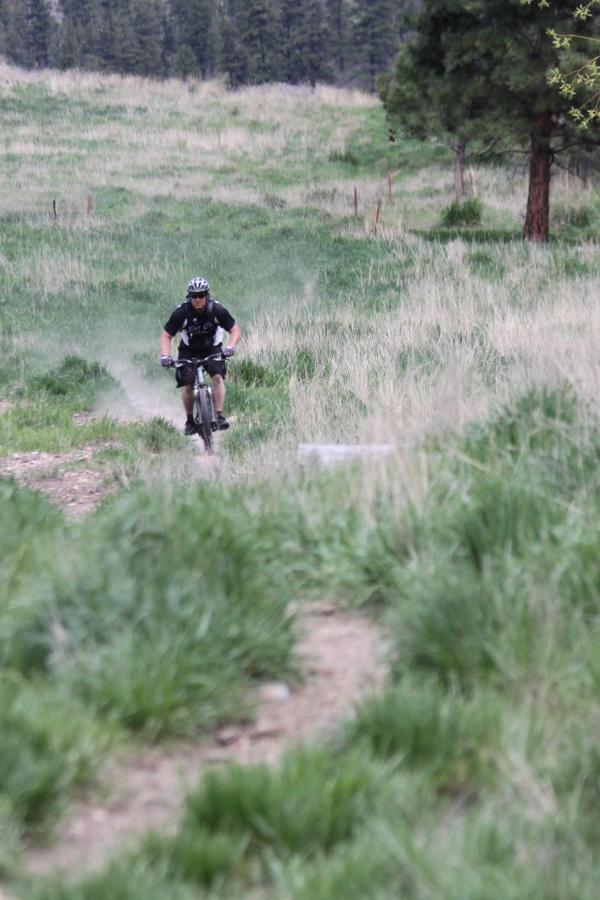 A mountain biker riding along a gravel path through a lush green landscape, kicking up dust as he pedals downhill. Tall grass surrounds the trail, and trees are visible in the background under a cloudy sky. Wagonhammer Trail System mountain bike trail.