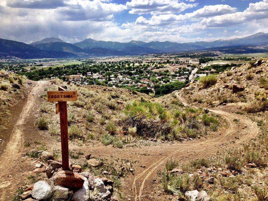 A dirt trail winding through hilly terrain, featuring a wooden sign labeled "FRONT SMO," overlooking a valley with a small town and mountains in the background. The sky is partly cloudy, casting shadows over the landscape. Frontside mountain bike trail.