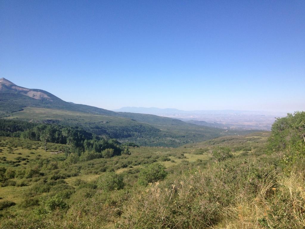 A scenic landscape view showcasing rolling green hills, dotted with trees, under a clear blue sky. In the background, a range of mountains can be seen, contributing to the picturesque natural setting. Hazard County mountain bike trail.