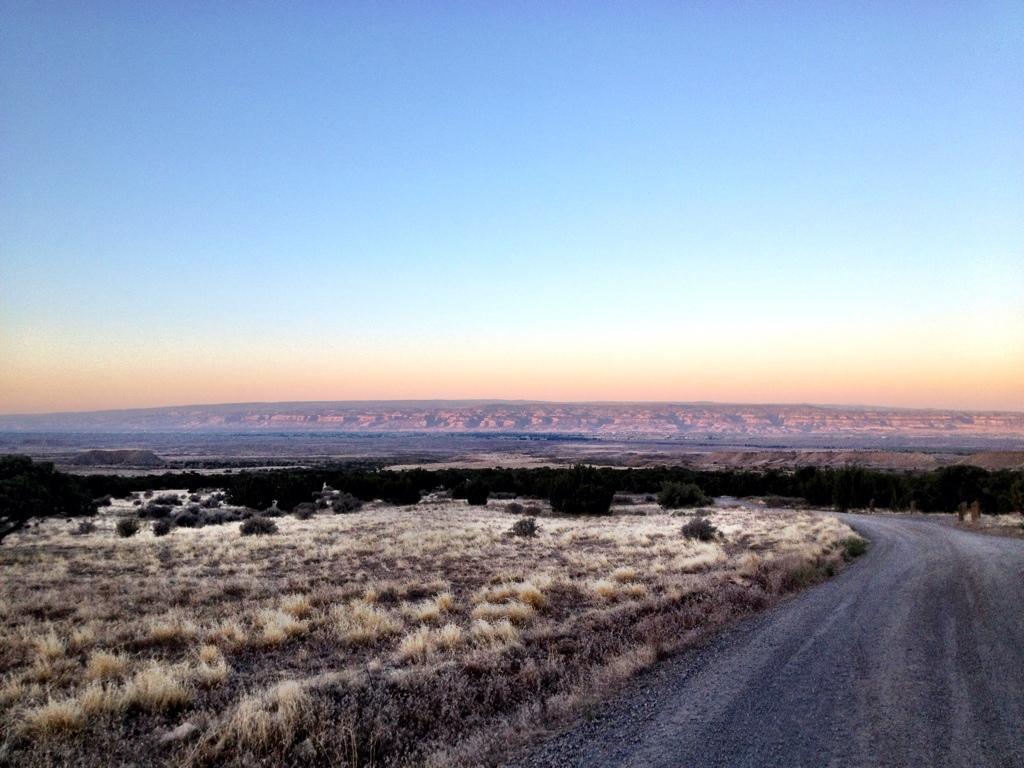 A serene landscape featuring a gentle gravel road winding through a grassy field, leading toward distant layered hills under a clear sky transitioning from soft orange to blue at dusk. Kessel Run mountain bike trail.
