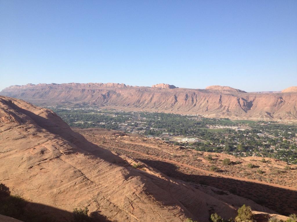 A panoramic view of a rocky landscape with layered red and brown cliffs in the background, contrasting with a green valley below. The sky is clear and blue, providing a bright backdrop to the natural scenery. The foreground features a rocky terrain with sparse vegetation. Slickrock mountain bike trail.