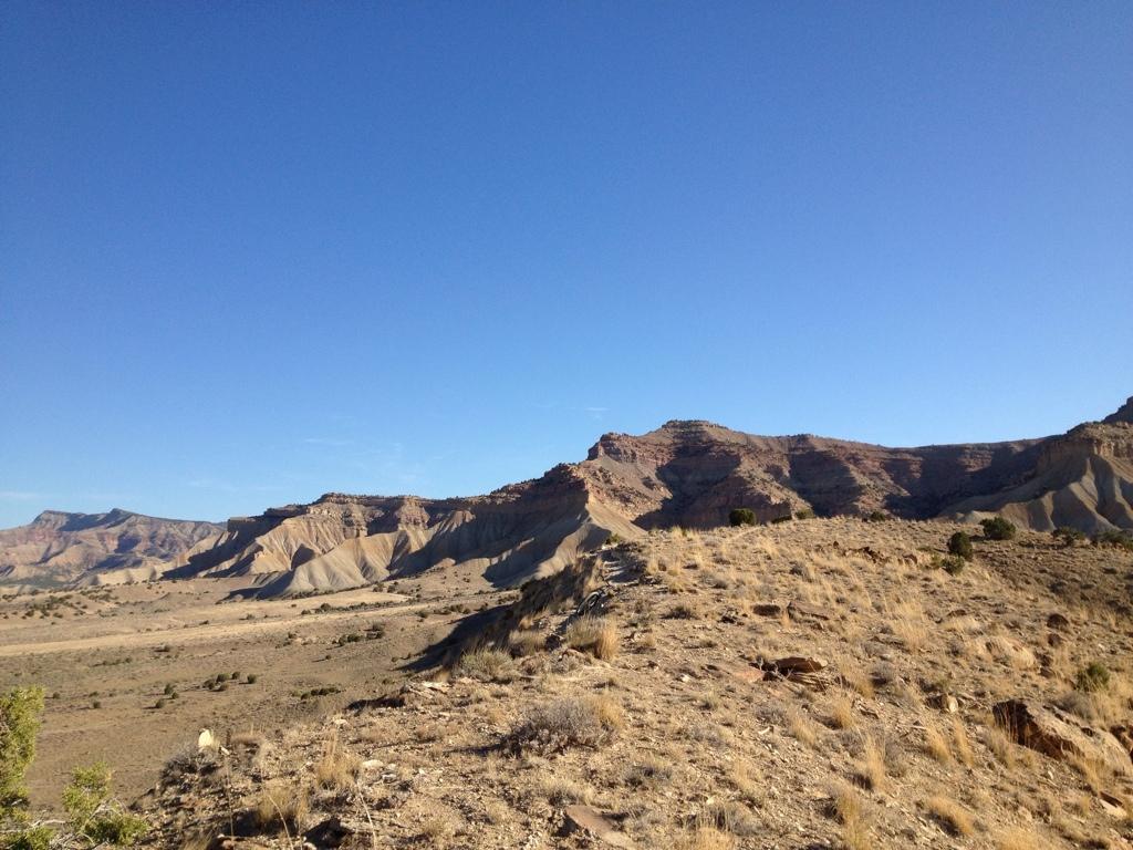 A panoramic view of a rugged desert landscape featuring layered hills and rocky formations under a clear blue sky. The foreground includes sparse vegetation and dry grass, while the distant mountains create a striking backdrop. Zippety Do Dah mountain bike trail.