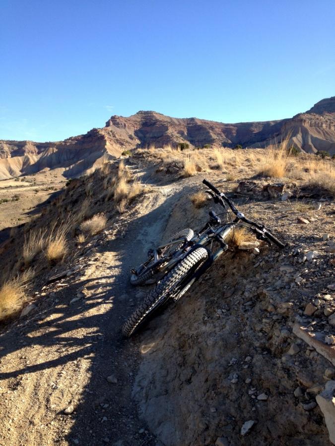 A mountain bike lying on its side on a rocky trail, surrounded by dry grass and rugged terrain, with scenic hills and blue sky in the background. Zippety Do Dah mountain bike trail.