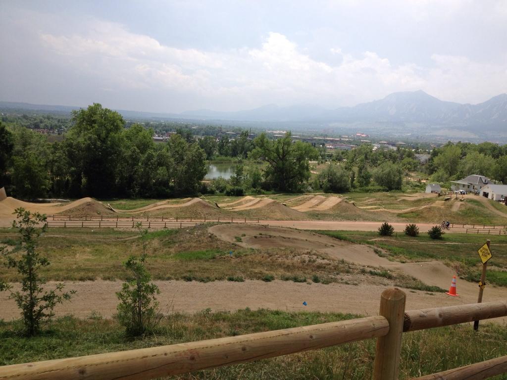A scenic view of a BMX bike park, featuring a dirt track with several jumps, surrounded by greenery and trees. In the background, a lake is visible along with a cityscape and distant mountains under a cloudy sky. A wooden fence is in the foreground, and a warning sign is posted nearby. Valmont Bike Park mountain bike trail.