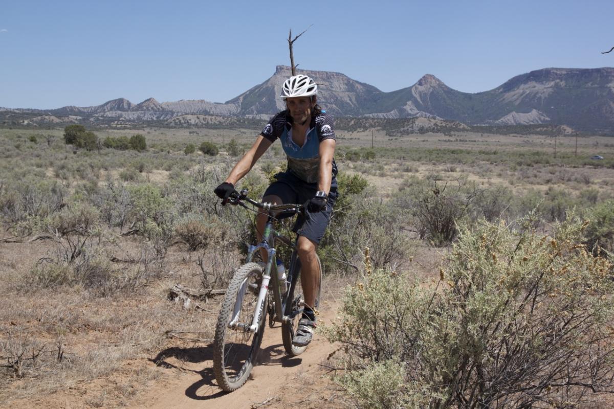 A person riding a mountain bike on a dirt trail surrounded by shrubs and open landscape, with rugged mountains in the background under a clear blue sky. Phil's World mountain bike trail.