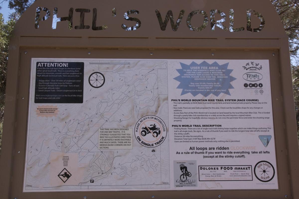 A signboard titled "Phil's World" displaying a detailed map and information about the Phil's World Mountain Bike Trail System. The sign includes important user instructions, a trail description, and safety guidelines. It outlines trail features, distances, and emphasizes that all loops should be ridden clockwise. Various logos and user fee information are also present on the sign. The background features natural scenery, indicating an outdoor setting. Phil's World mountain bike trail.