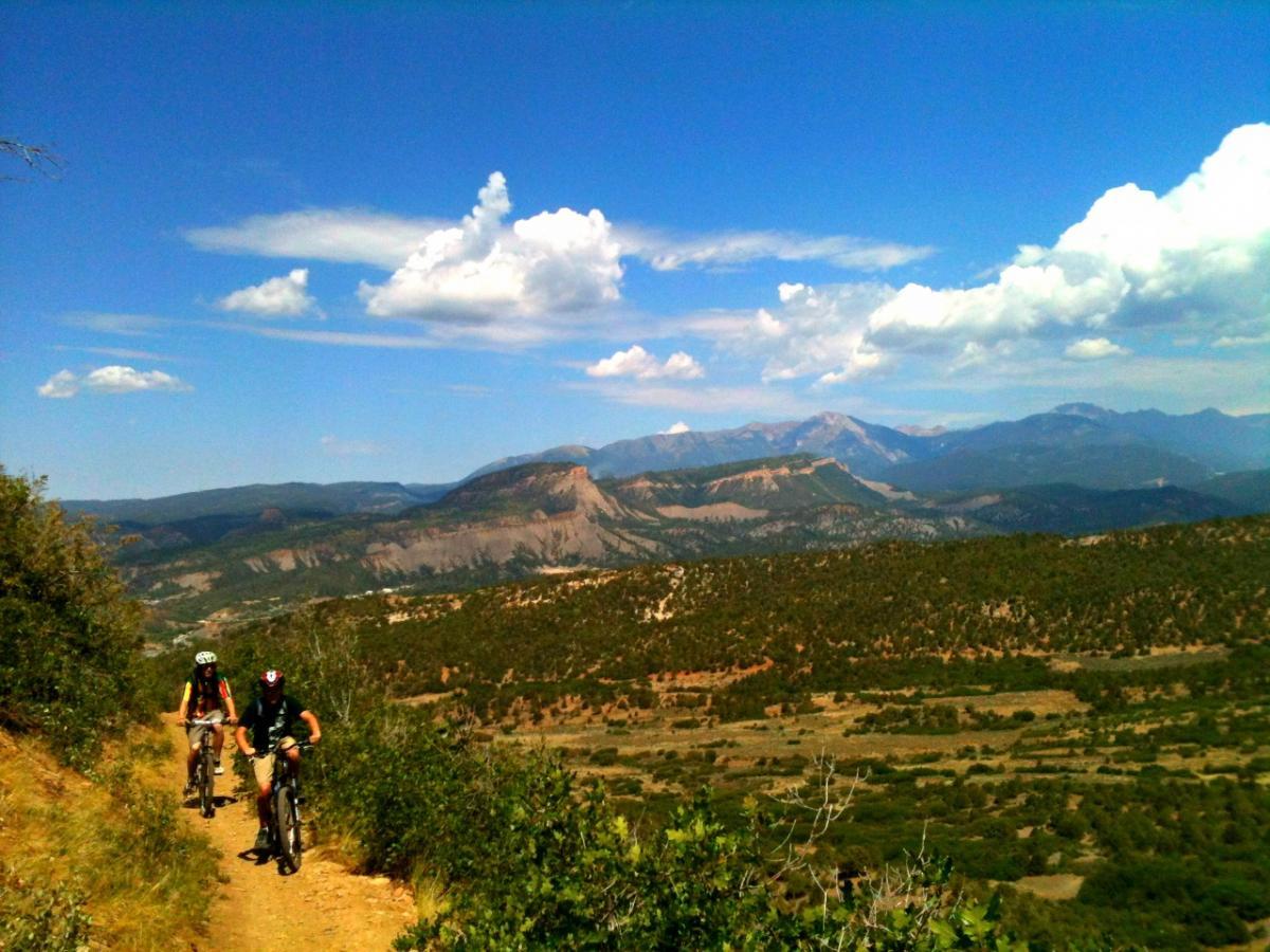 Two mountain bikers navigate a dirt trail through a scenic landscape featuring rolling hills and distant mountains under a blue sky with scattered clouds. Green vegetation lines the path, highlighting the natural beauty of the setting. Horse Gulch mountain bike trail.