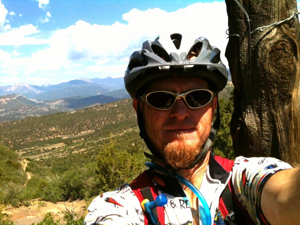 A person in a cycling helmet and sunglasses poses for a selfie against a backdrop of mountains and a blue sky. The landscape features greenery and rolling hills, suggesting an outdoor adventure. The individual is wearing a colorful cycling jersey and appears to be enjoying a sunny day. Horse Gulch mountain bike trail.