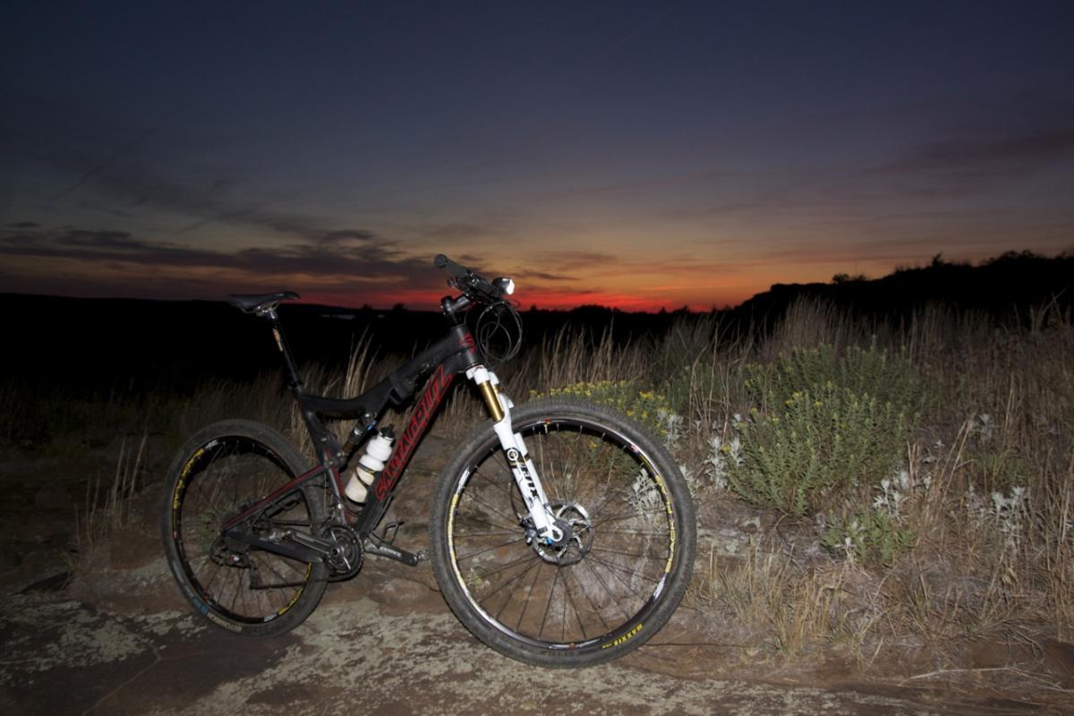 A mountain bike parked on rocky terrain during sunset, with vibrant orange, pink, and purple skies in the background. The bike features a black frame with red accents and is equipped with a front light. Surrounding the bike are tall grasses and small bushes, suggesting a natural outdoor setting. Switchgrass mountain bike trail.