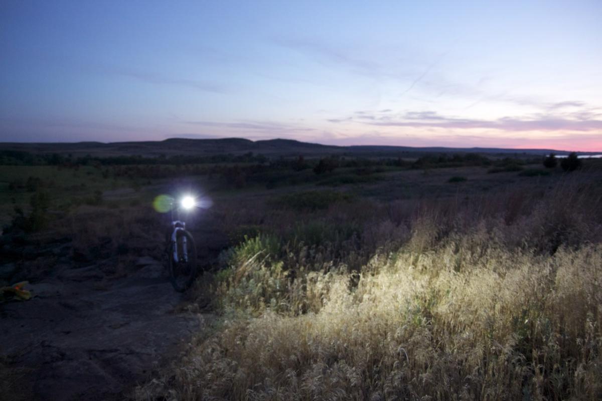 A mountain bike is parked on a rocky path at dusk, with its front light glowing brightly. In the background, a vast, open landscape stretches out, featuring rolling hills and a colorful sunset sky. Grassy terrain and scattered shrubs surround the bike, adding to the serene outdoor atmosphere. Switchgrass mountain bike trail.