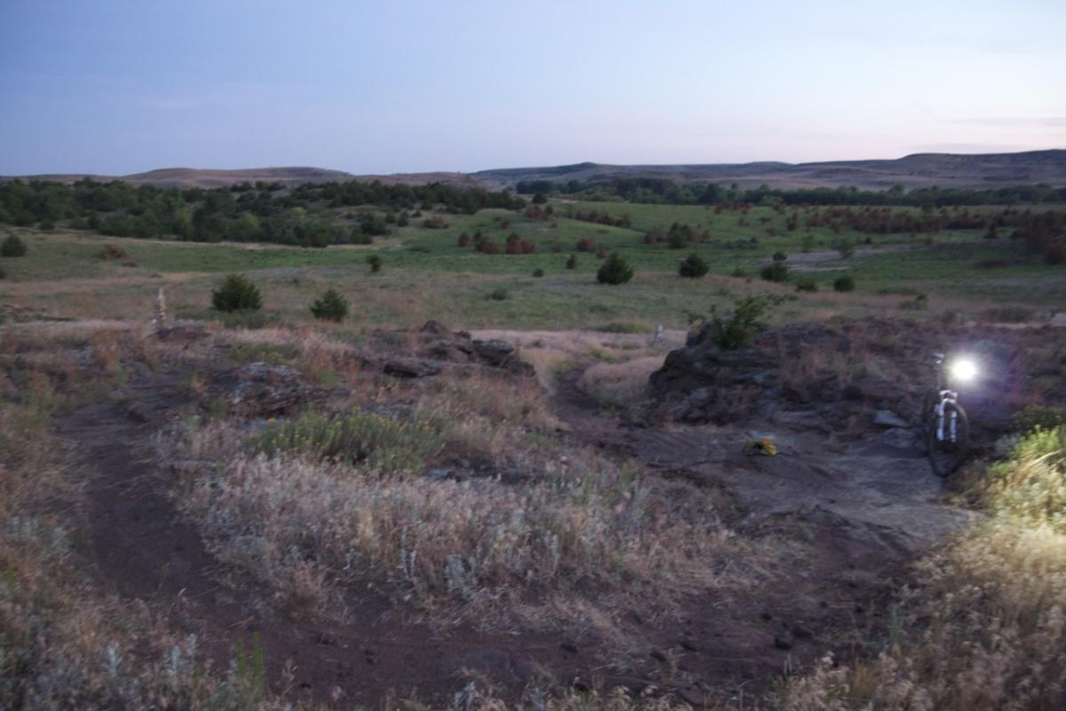 A twilight landscape featuring rolling hills and grassy fields, with patches of trees scattered throughout. A rocky outcrop is visible in the foreground, alongside a mountain bike illuminated by a light. The scene captures a serene, natural setting just before nightfall. Switchgrass mountain bike trail.