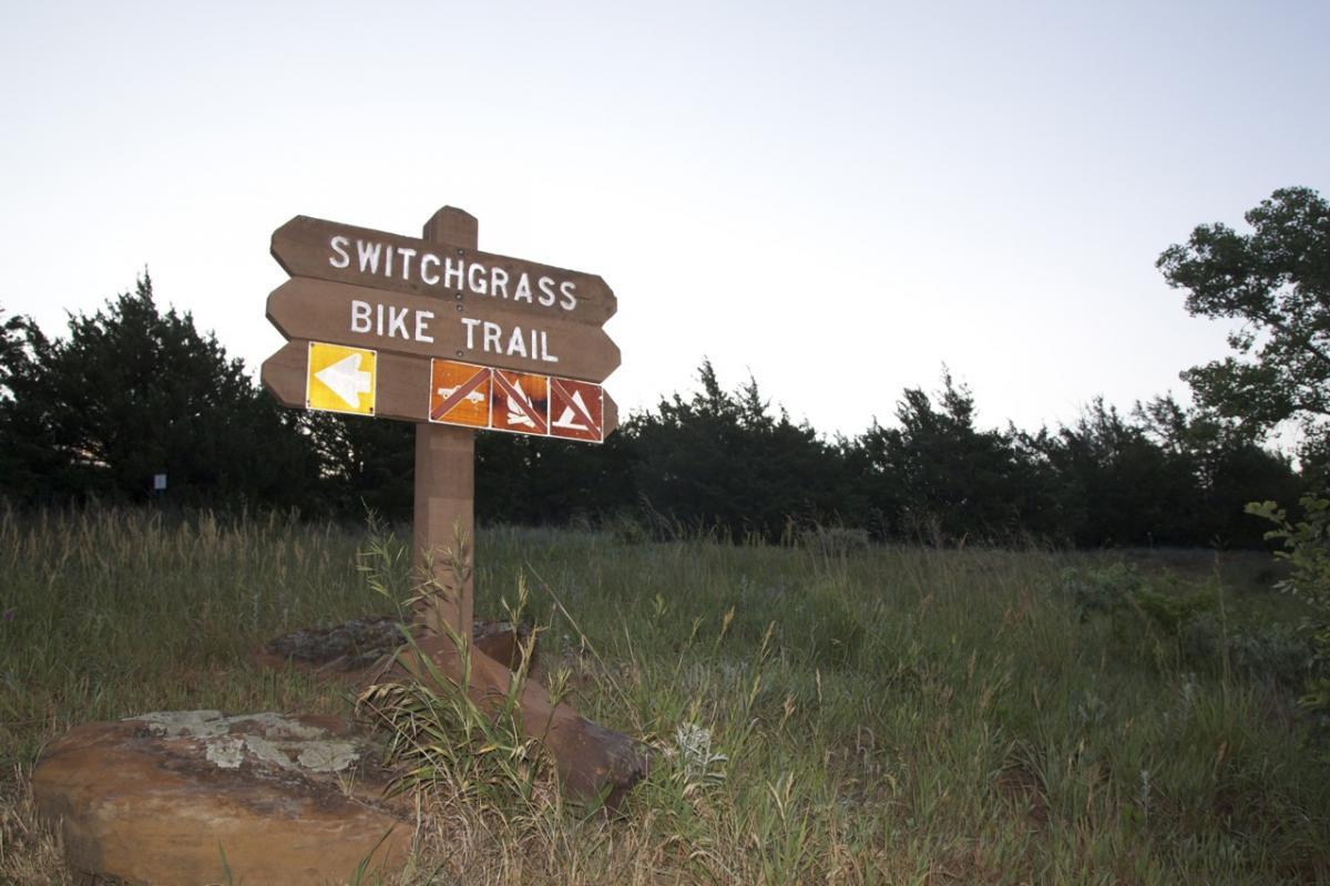 A wooden signpost displaying directions for the Switchgrass Bike Trail, with arrows indicating various routes. The sign is surrounded by tall grass and trees, set against a twilight sky. Switchgrass mountain bike trail.