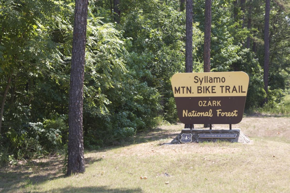 A sign for the Syllamo Mountain Bike Trail located in the Ozark National Forest, surrounded by lush green trees and foliage. The sign features a yellow and brown design with the trail name prominently displayed. Syllamo Trails mountain bike trail.