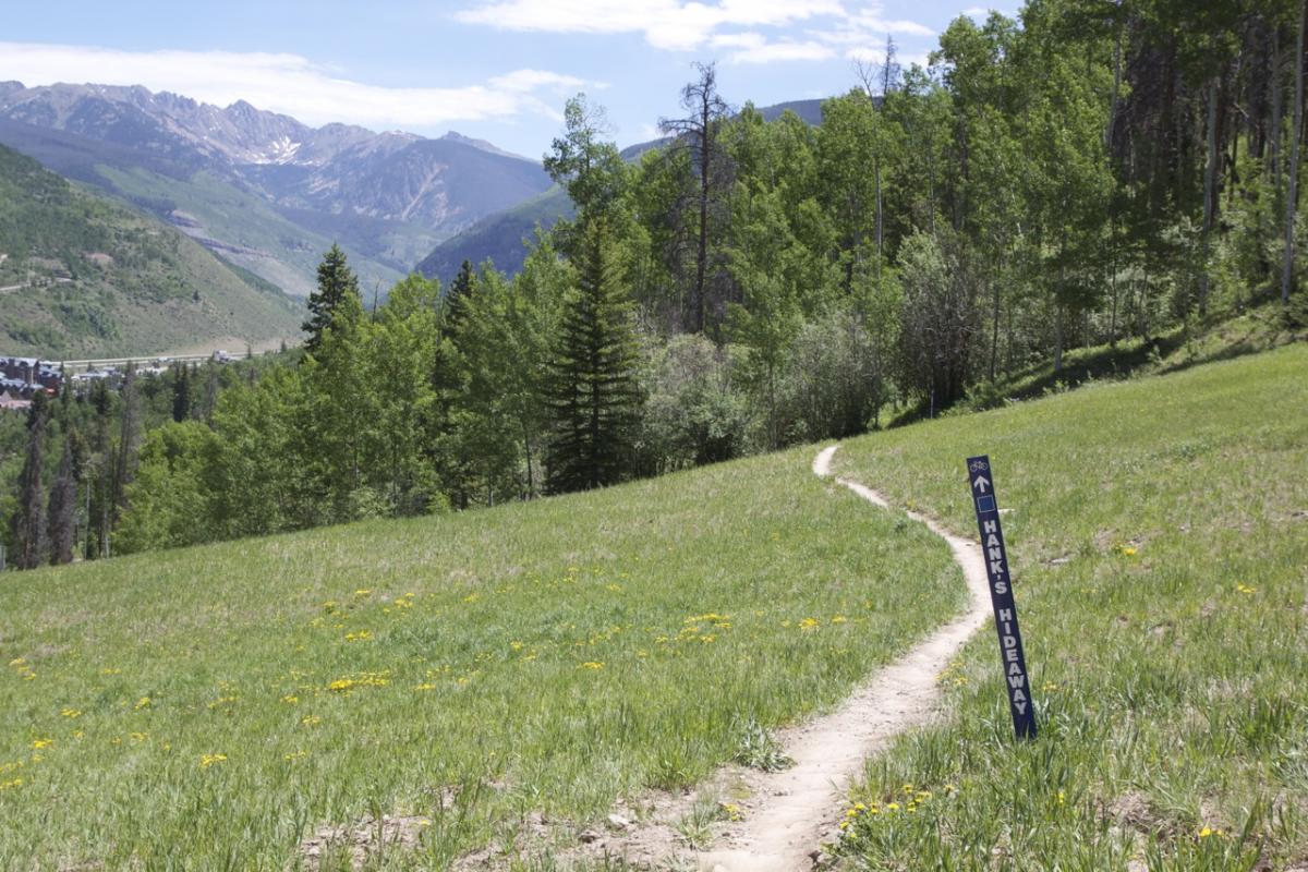 A dirt path winding through a grassy field, surrounded by green trees, with distant mountains in the background under a partly cloudy sky. A trail sign labeled "Hank's Hideaway" stands alongside the path. Vail Mountain Bike Park mountain bike trail.
