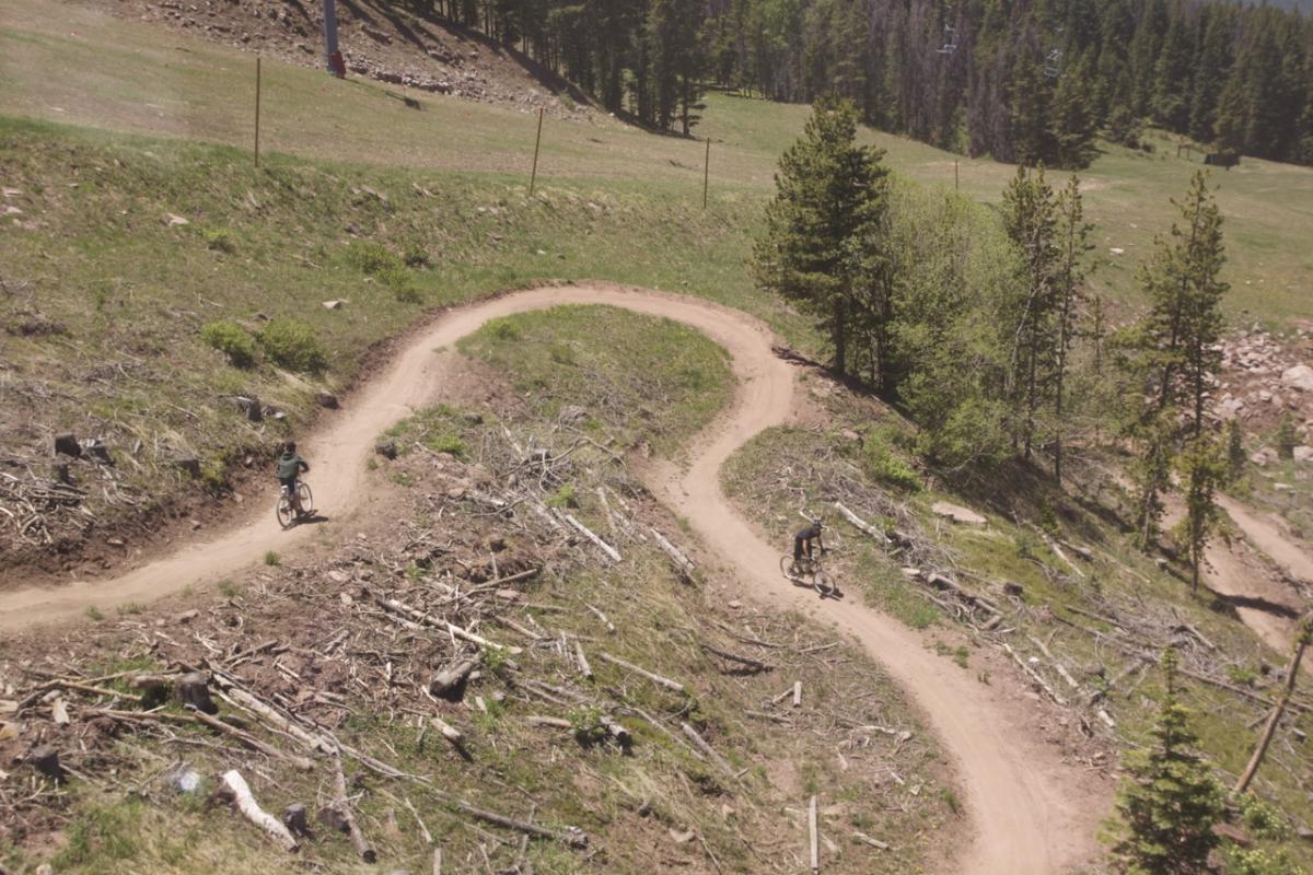 Aerial view of two mountain bikers riding on a winding dirt trail through a forested area, with patches of cleared land and scattered logs visible. Lush greenery surrounds the trail, and there are trees in the background. Vail Mountain Bike Park mountain bike trail.