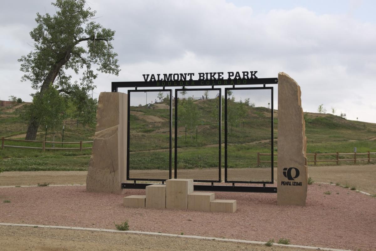 Entrance sign to Valmont Bike Park in Boulder, Colorado, featuring a black metal frame with the park's name and location. Two large stone pillars flank the entrance, with a logo at the base. The background includes grassy hills and trees under a cloudy sky. Valmont Bike Park mountain bike trail.
