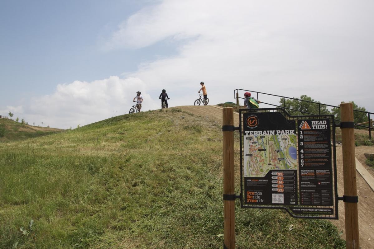 A group of children on bicycles at a terrain park, gathered on a grassy hill with a sign displaying park rules and a map. The sky is partly cloudy, and the area is surrounded by trees and hills. Valmont Bike Park mountain bike trail.