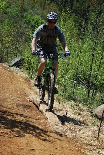 A cyclist navigating a narrow dirt trail on a green mountain bike, wearing a helmet and gloves, surrounded by trees and greenery in a forested area. Rocky Knob Park mountain bike trail.
